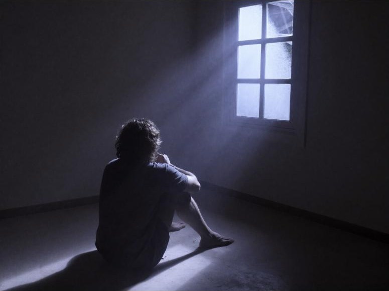 A man sitting alone on the floor of a dark room, lit by one window